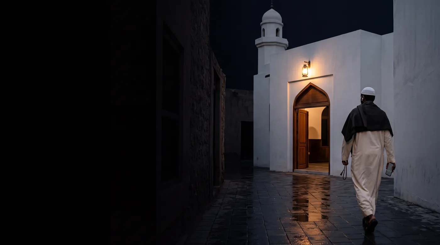A man in a kufi and thobe walking towards a small whitewashed mosque before dawn, his phone at his side showing a Madinah page in Criterion's Night mode.